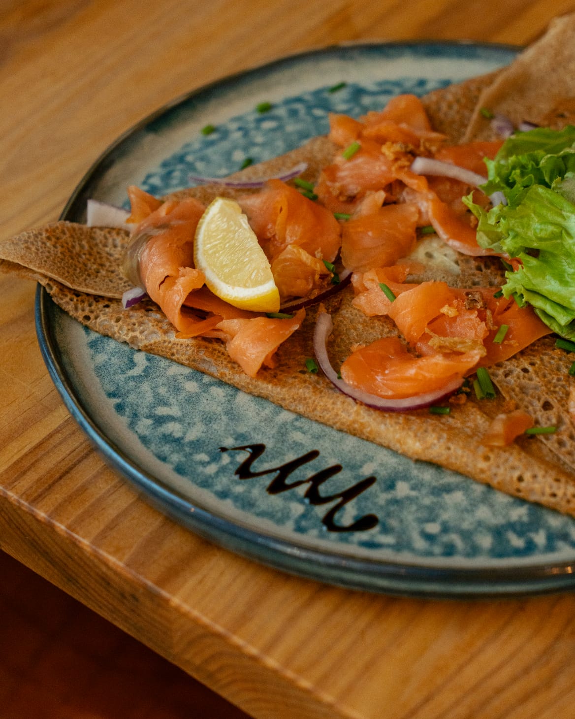 Galette de sarrasin au saumon fumé, oignon rouge, ciboulette et quartier de citron, servie avec de la salade sur une assiette signée Miam