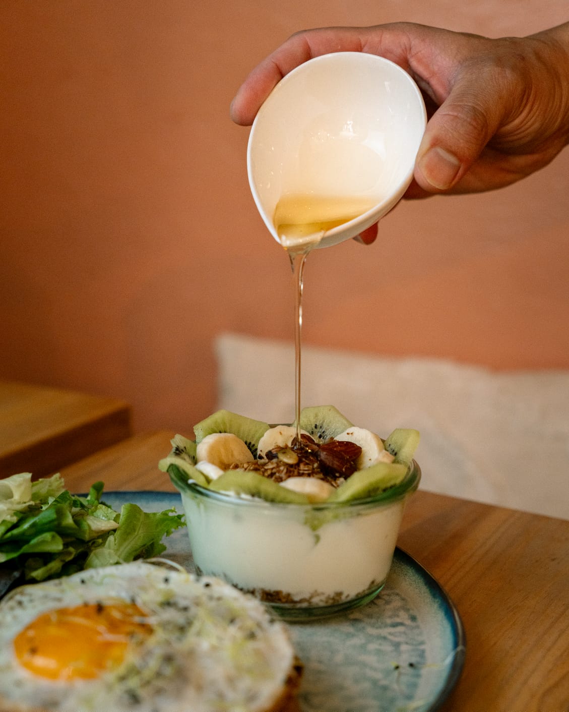 Filet de miel versé sur un Grano'miam : fromage blanc, granola maison, kiwi, banane et amandes, servi à côté d'un œuf au plat