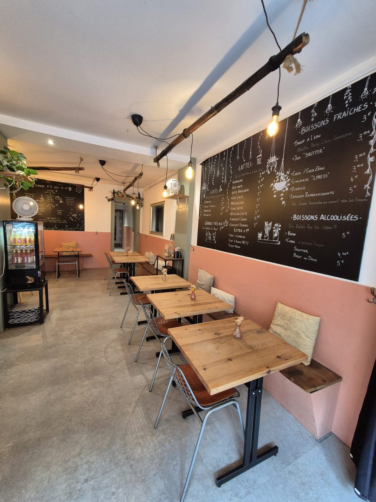 La salle du café : tables en bois et banquettes le long du mur pêche, sous les ardoises de la carte des boissons